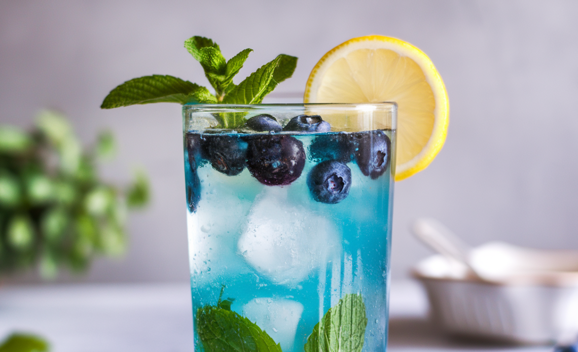 A glass of homemade blueberry mint lemonade, garnished with fresh blueberries and mint leaves, served over ice with a lemon slice on the rim.